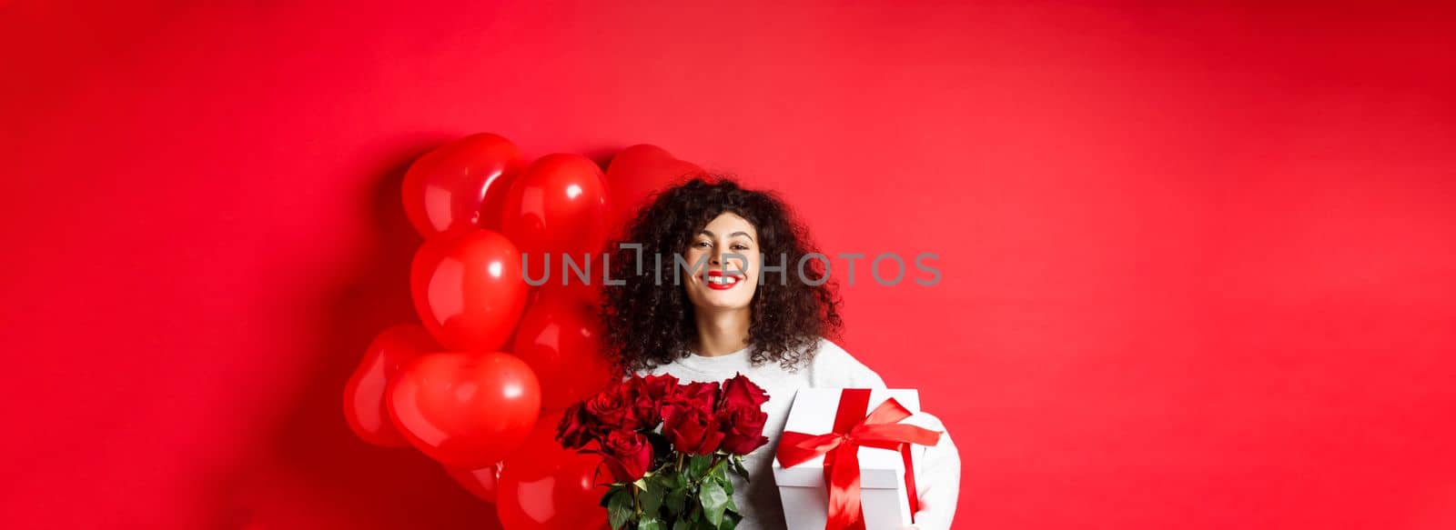 Smiling happy woman holding box with gift and red roses from boyfriend, celebrating Valentines day, standing near romantic hearts balloons, standing over studio background.