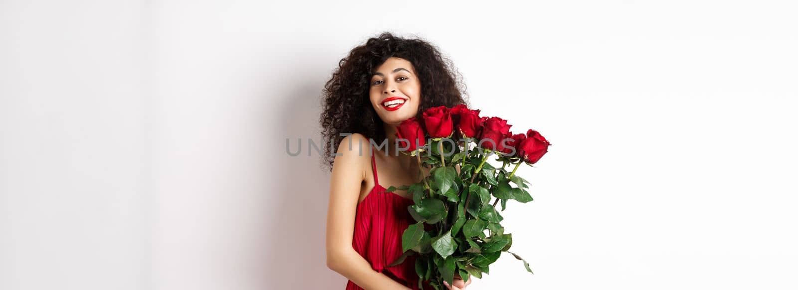 Romantic lady with curly hair and fashionable dress, holding bouquet of red roses and smiling, standing happy on white background.