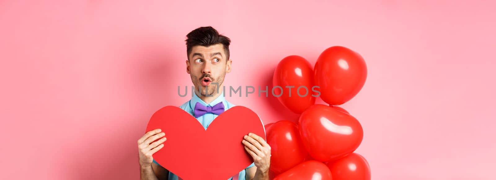 Valentines day concept. Cute young man looking left amused, showing red heart cutout and standing near balloons, pink background by Benzoix