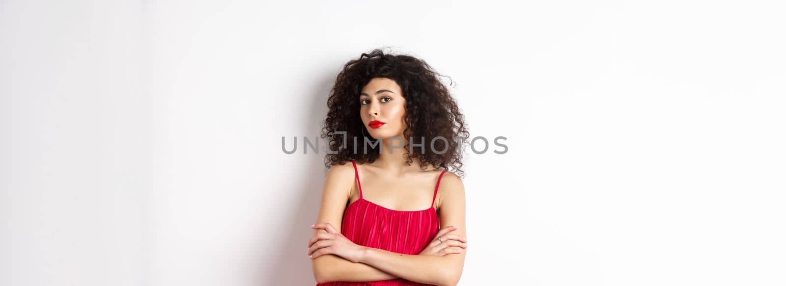 Sassy woman with curly hairstyle, wearing red dress and makeup, cross arms on chest, standing over white background.