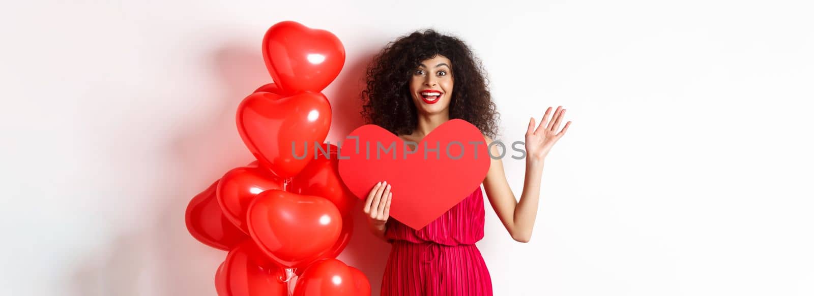 Valentines day and love concept. Cheerful young woman in elegant red dress, standing near romantic balloons and holding big red heart cutout, waving hand to say hi, waiting for date by Benzoix