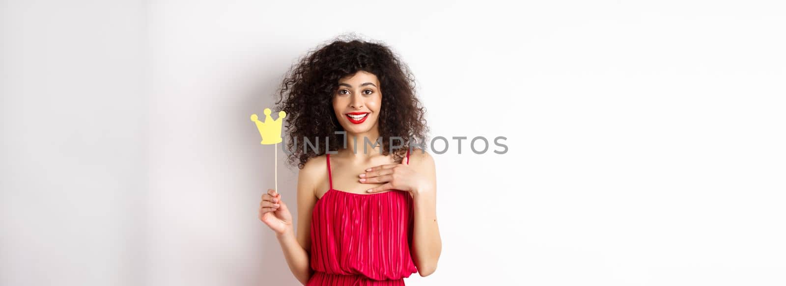 Confident pretty lady in red dress, holding queen crown on stick and looking excited, standing on white background.