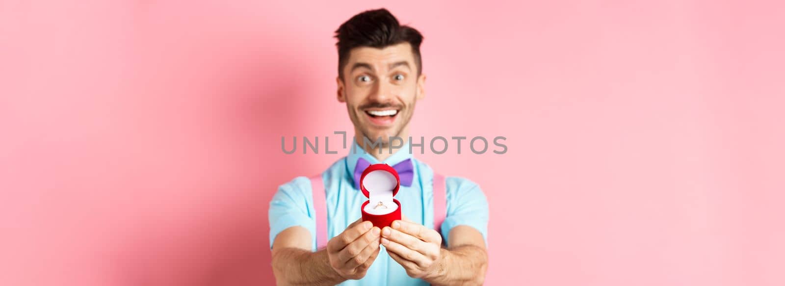 Valentines day. Romantic young man in bow-tie stretch hands with engagement ring and smiling, asking to marry him, making proposal, standing over pink background by Benzoix