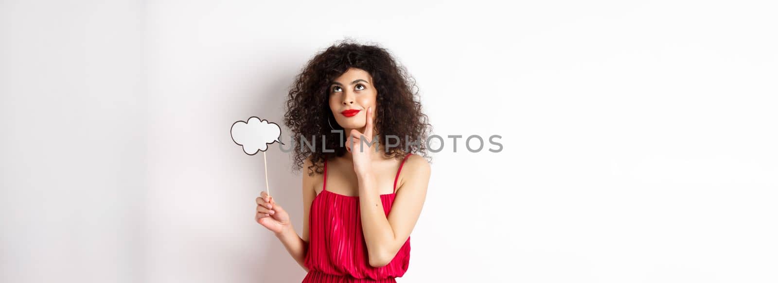 Thoughtful girl in red dress, holding small cloud and looking up with pleased smile, imaging things, standing over white background.