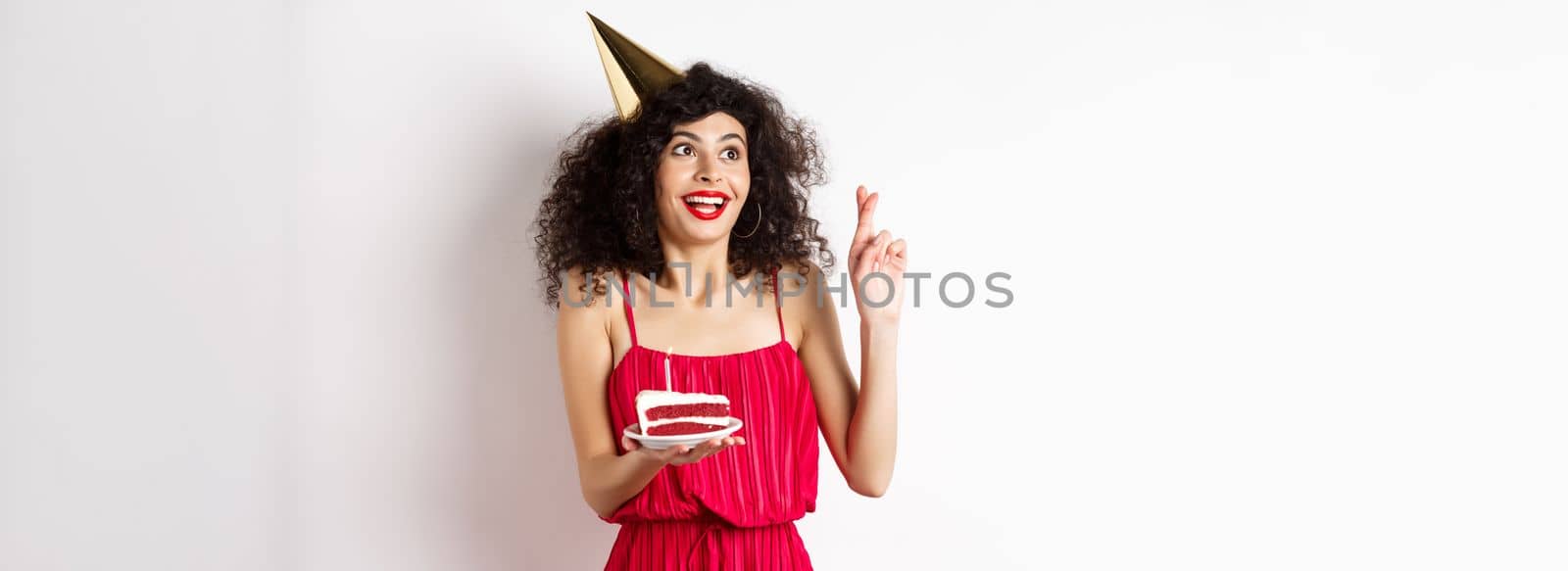 Excited birthday girl making wish, cross fingers good luck and looking aside at logo, celebrating bday, holding piece of cake, white background.