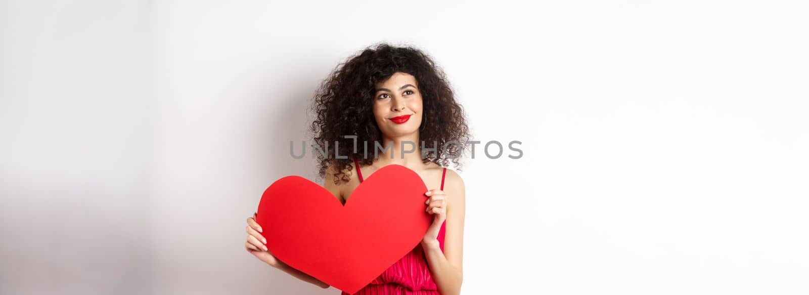 Young romantic woman dreaming about love on Valentines day, looking for soulmate, holding big red heart cutout and gazing pensive left, white background.