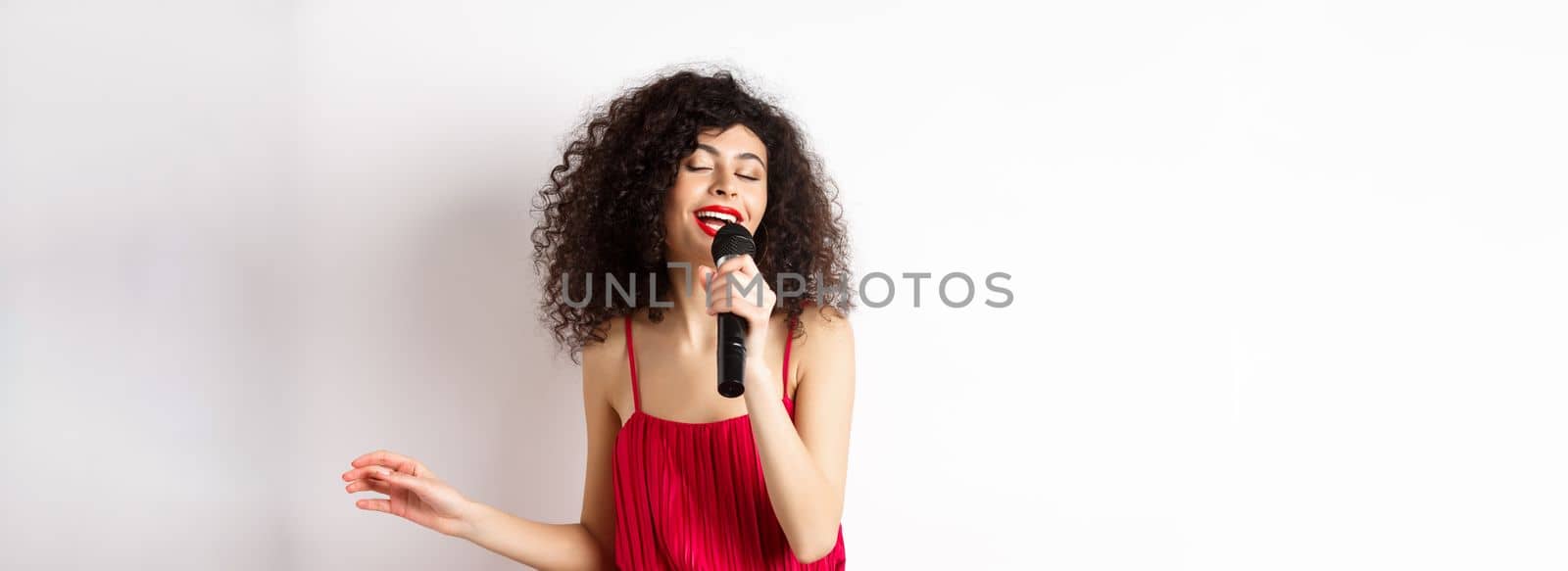 Happy elegant woman in red dress performing with microphone, singing karaoke and smiling, standing over white background.