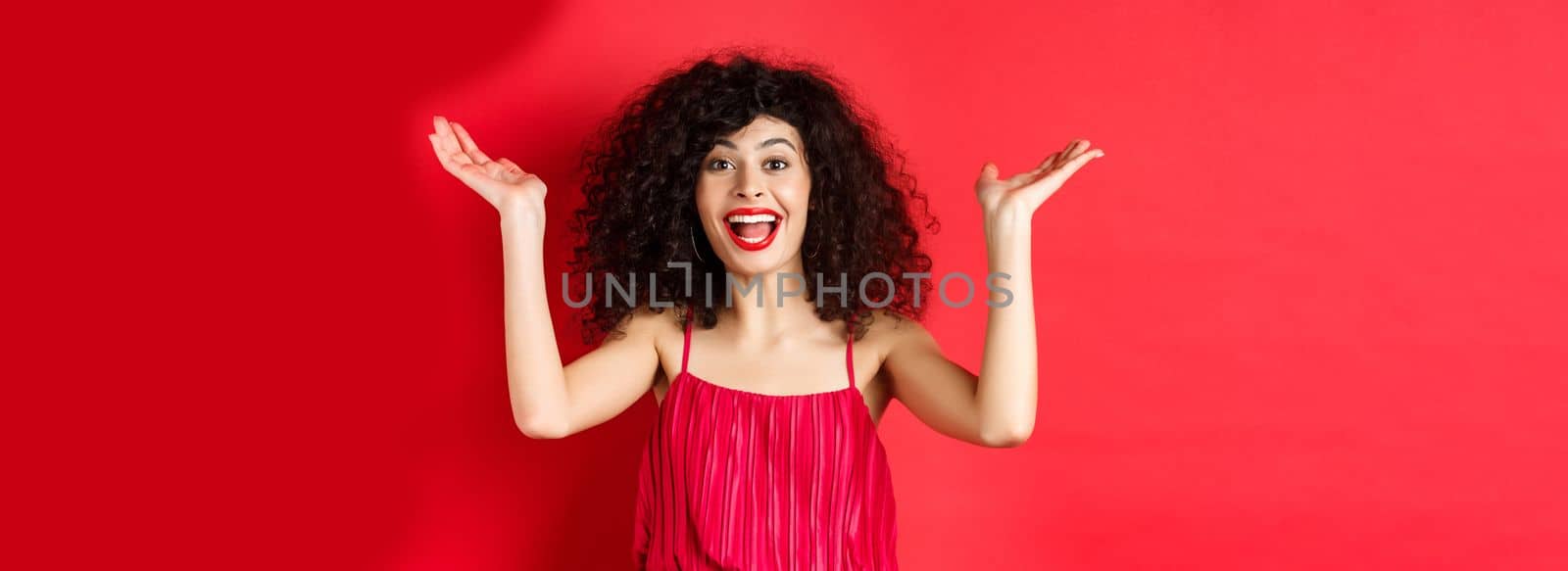 Excited beautiful woman raising hands up and scream with happiness and joy, applause with rejoice, standing on red background by Benzoix