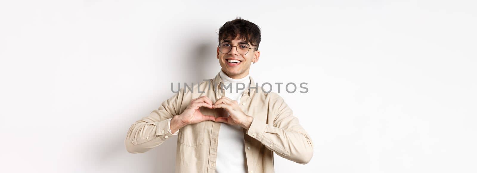 Happy Valentines. Handsome young man in glasses say I love you, showing heart sign and smiling at girlfriend, celebrating lovers day, standing over white background by Benzoix