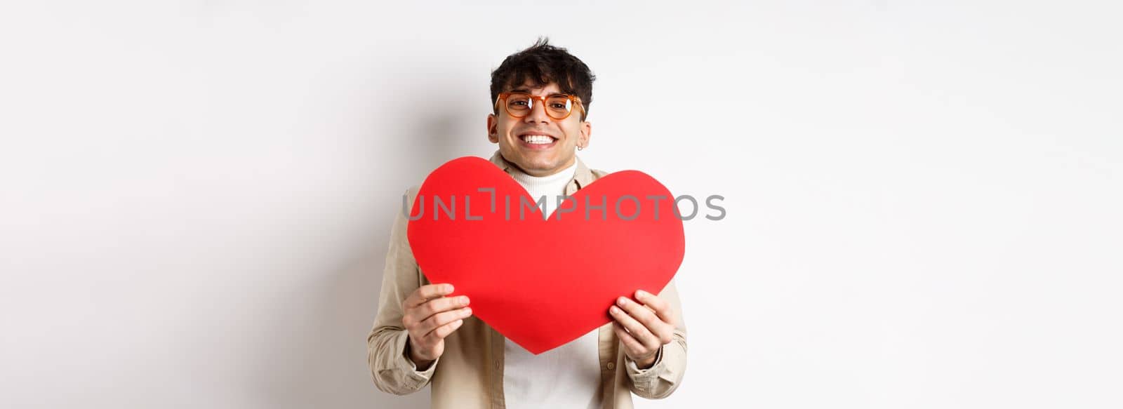 Cheerful young man smiling and looking thrilled at camera, showing big red heart cutout on Valentines day, making romantic gift to lover, white background by Benzoix