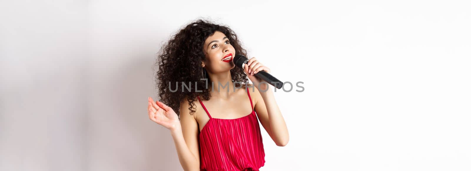 Beautiful lady in red dress singing songs in microphone, smiling and looking up, standing on white background.