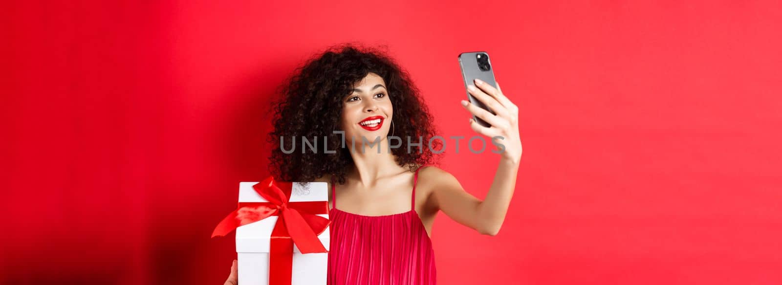 Beautiful girlfriend with curly hair, wearing evening dress, taking selfie with gift from lover, photographing on smartphone and smiling, standing over red background.
