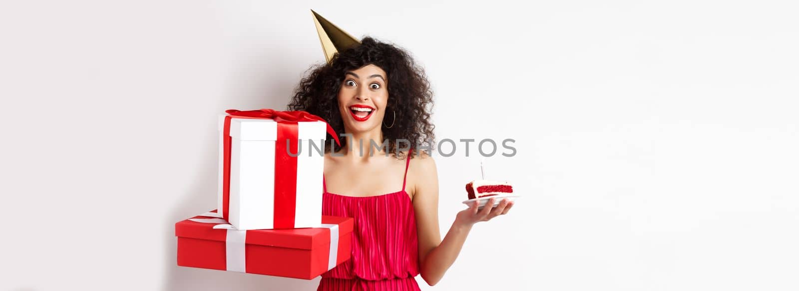 Cheerful girl in party hat and red dress, celebrating birthday, holding surprise gift and b-day cake, enjoying holiday, standing over white background by Benzoix