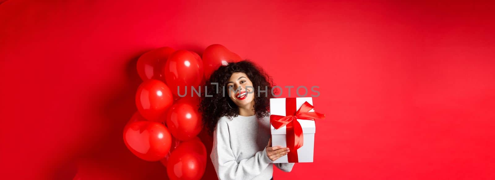 Holidays and celebration. Happy birthday girl holding gift and posing near party helium balloons, smiling excited at camera, red background.