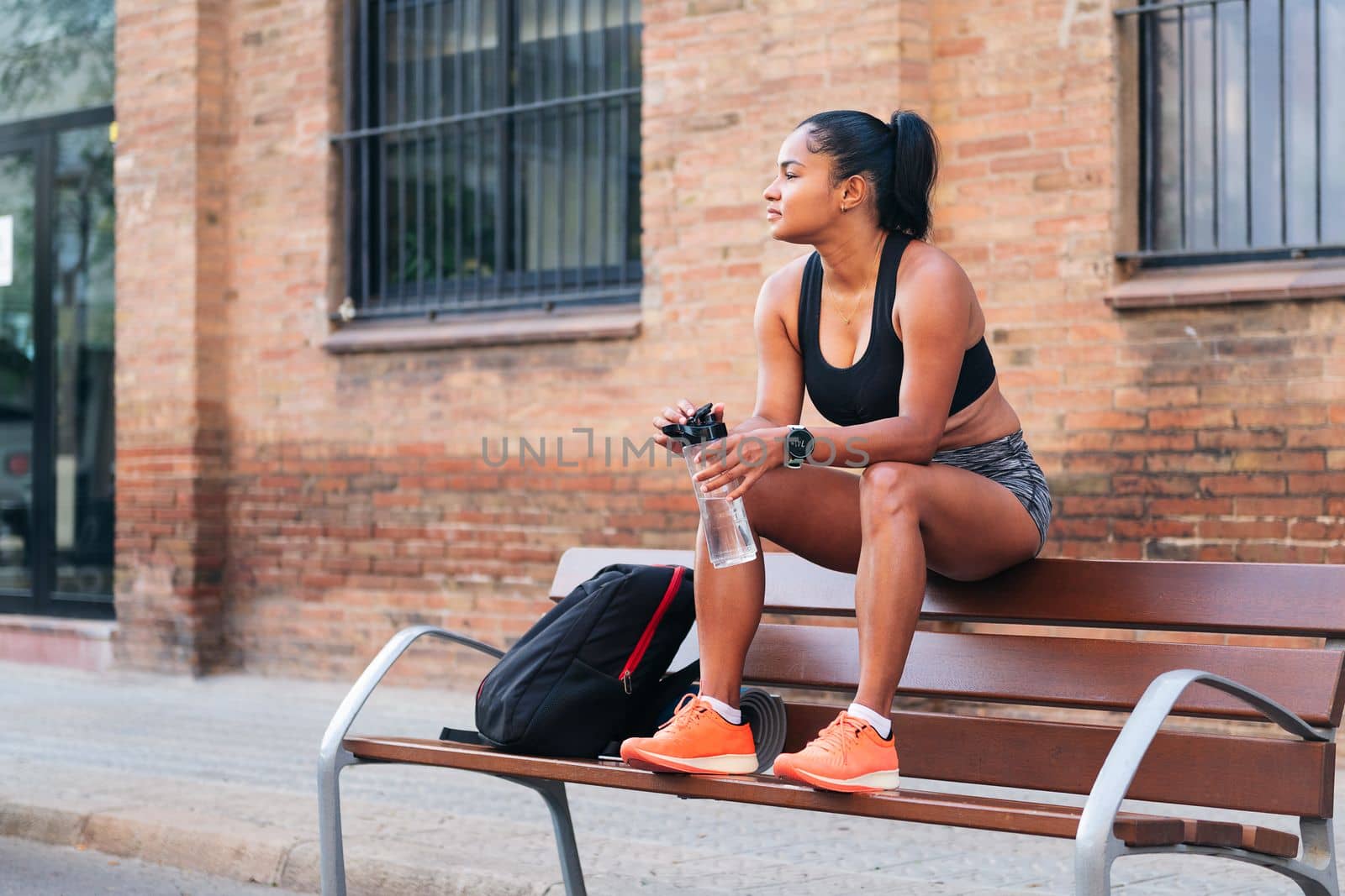 latina female athlete with a bottle of water resting while sitting on a bench during a break in her workout, urban sport and healthy lifestyle concept, copy space for text