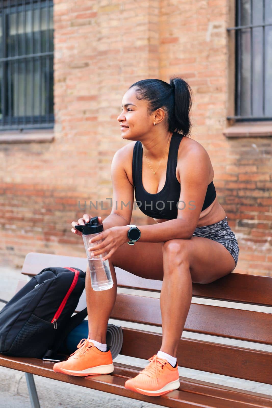 latina female athlete with a bottle of water resting while sitting on a bench during a break in her workout, urban sport and healthy lifestyle concept, copy space for text