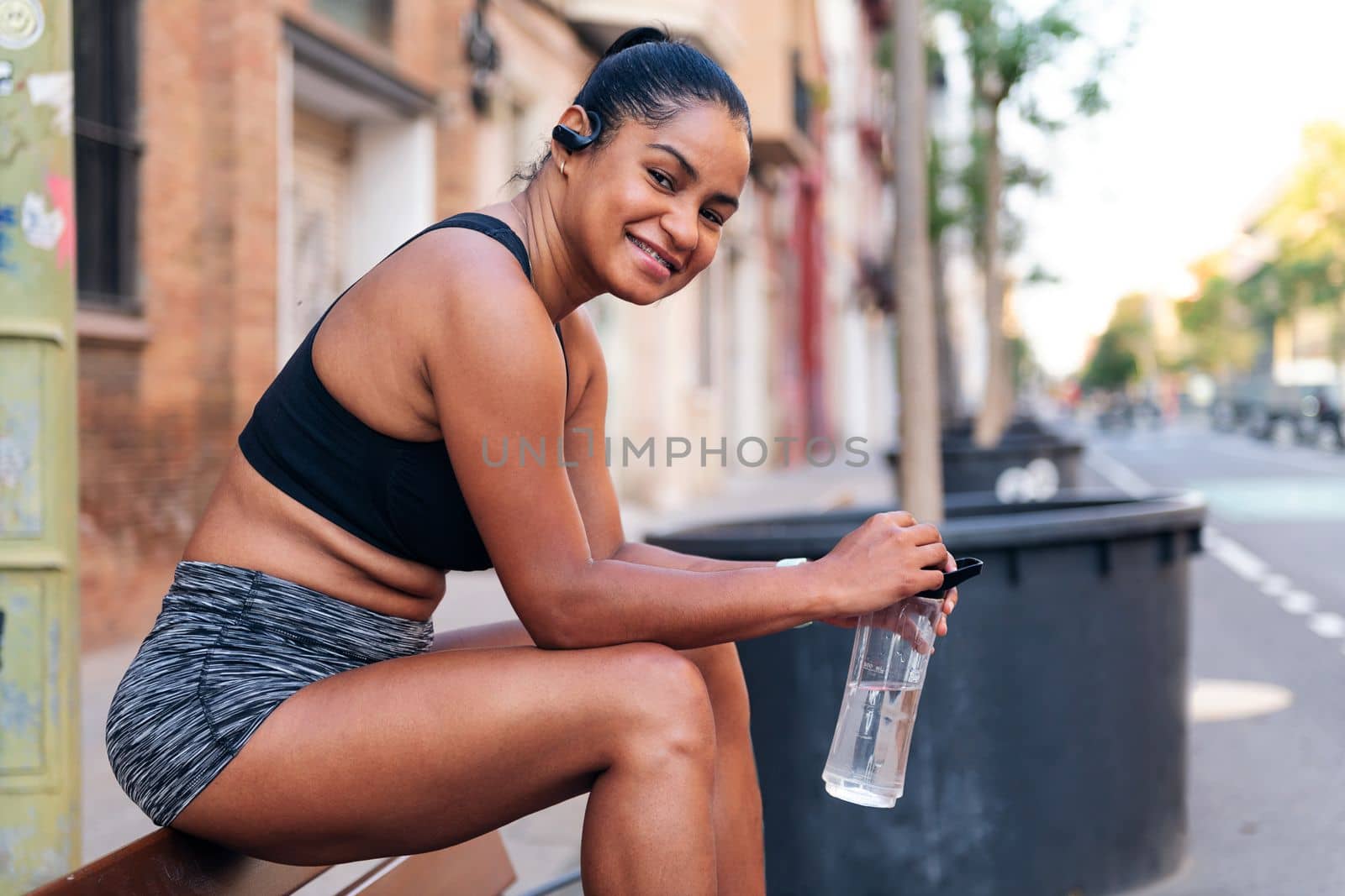 latina sportswoman holding a water bottle smiles while sitting looking at camera during a break in her workout, urban sport and healthy lifestyle concept, copy space for text