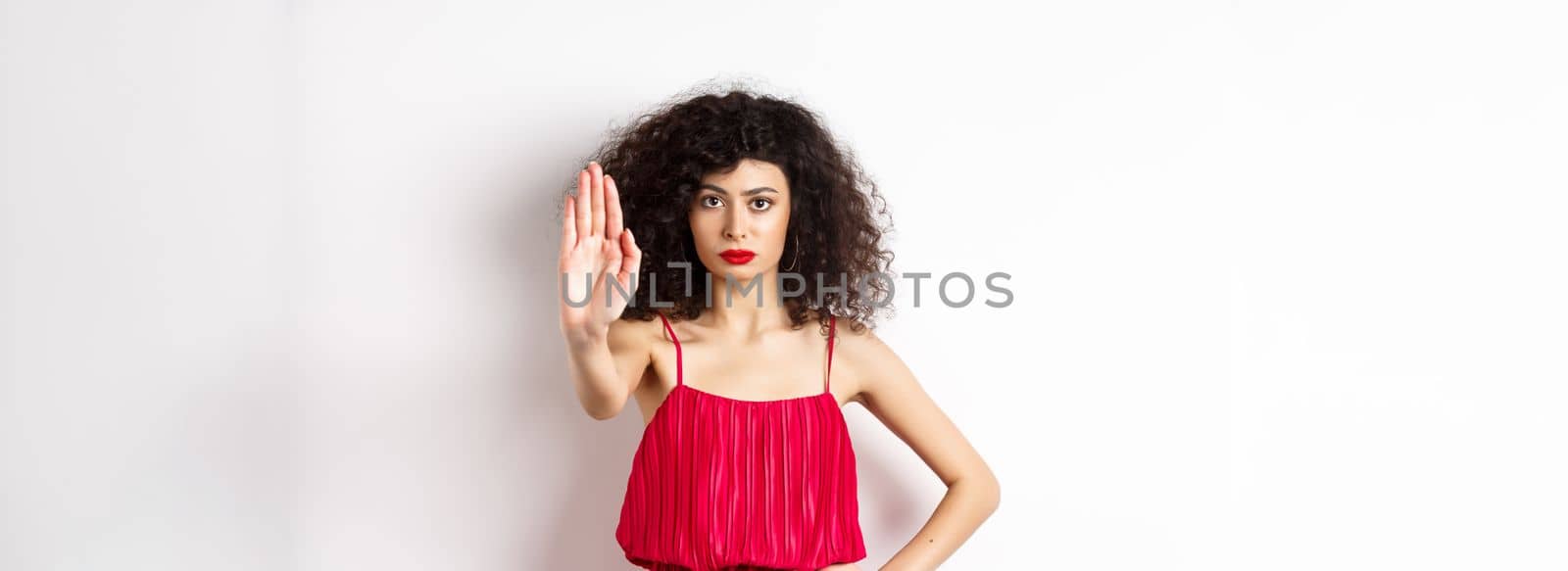 Serious and confident woman in red dress and makeup stretch out hand, tell to stop, prohibit and forbid something, standing over white background by Benzoix