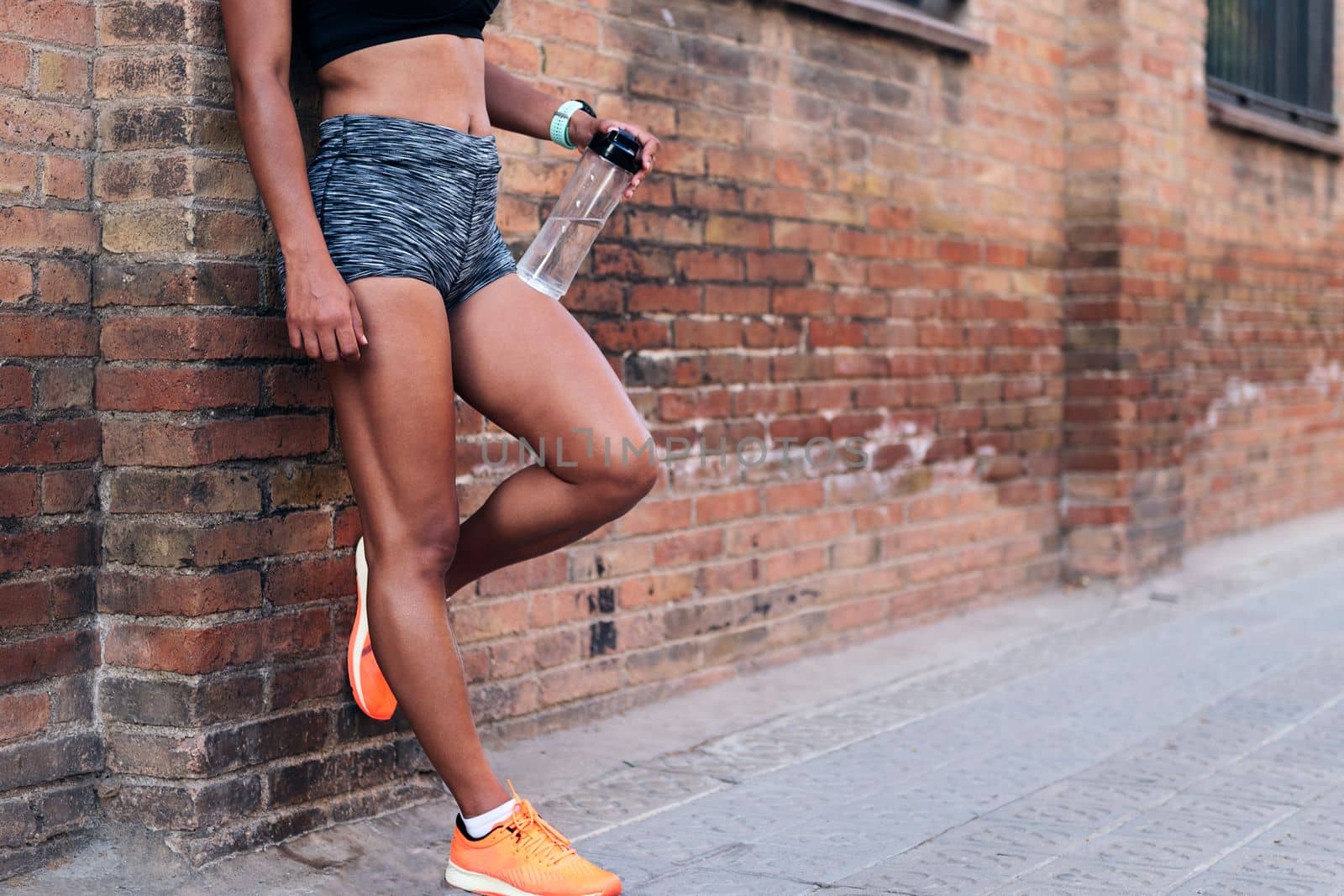 body of an unrecognizable sportswoman holding a bottle of water while resting against a brick wall during a break in her training, urban sport and healthy lifestyle concept, copy space for text