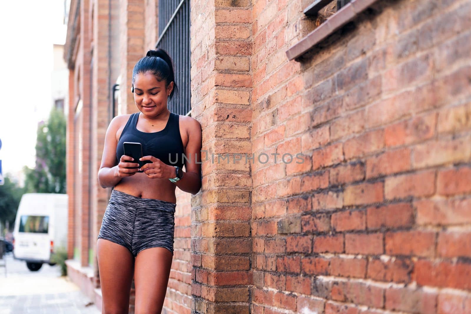 latin sportswoman using her mobile phone resting against a brick wall during a break in her workout, urban sport and healthy lifestyle concept, copy space for text