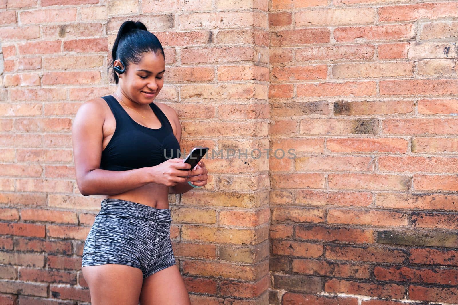 latin sportswoman using her mobile phone resting against a brick wall during a break in her workout, urban sport and healthy lifestyle concept, copy space for text