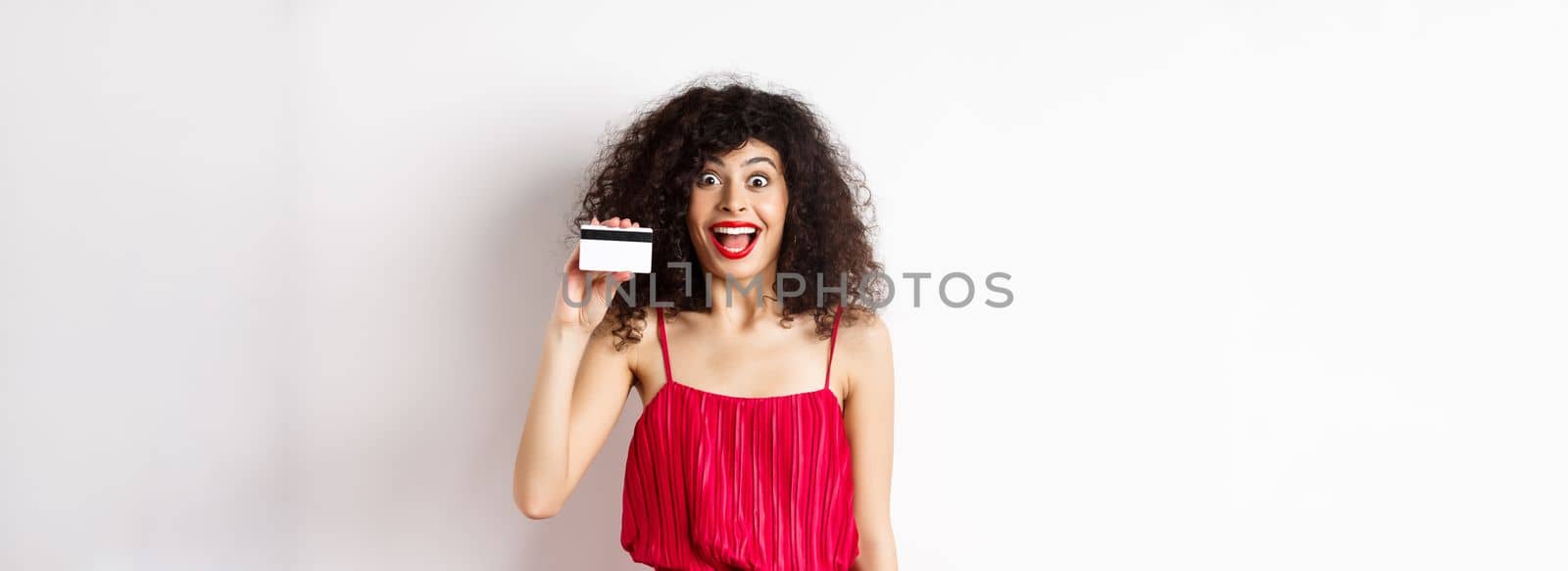 Excited smiling woman in red dress, showing plastic credit card and looking happy, standing over white background by Benzoix