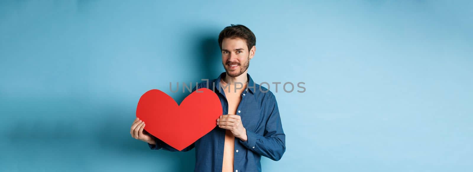 Happy man showing valentines heart and smiling, make romantic gift on lovers day, standing over blue background by Benzoix