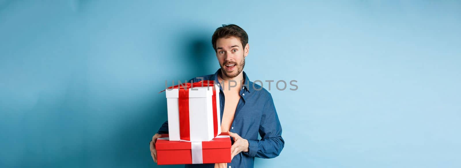 Handsome bearded guy holding Valentines day gifts for lover, standing with presents in boxes and looking at camera, blue background.