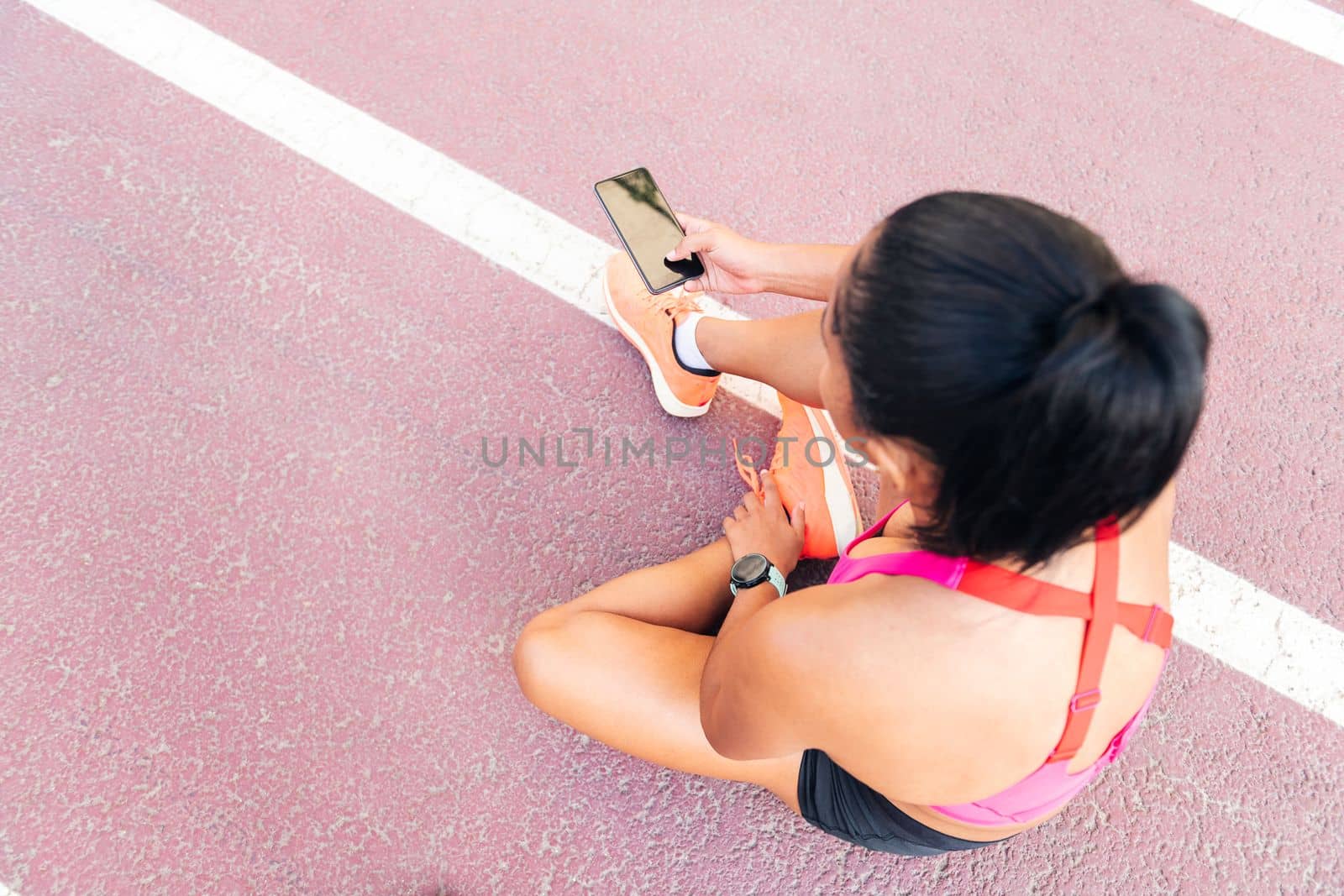 top view of an unrecognizable female athlete using her mobile phone sitting on the athletics track after her workout, concept of sport and healthy lifestyle, copy space for text