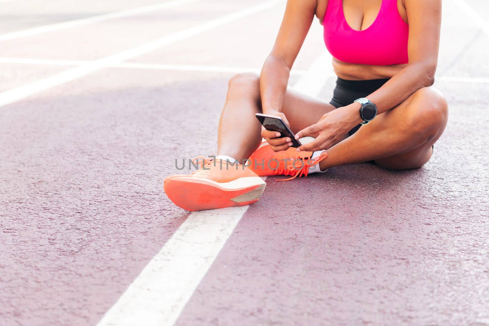 unrecognizable female athlete using her mobile phone sitting on the athletics track after her workout, concept of sport and healthy lifestyle, copy space for text