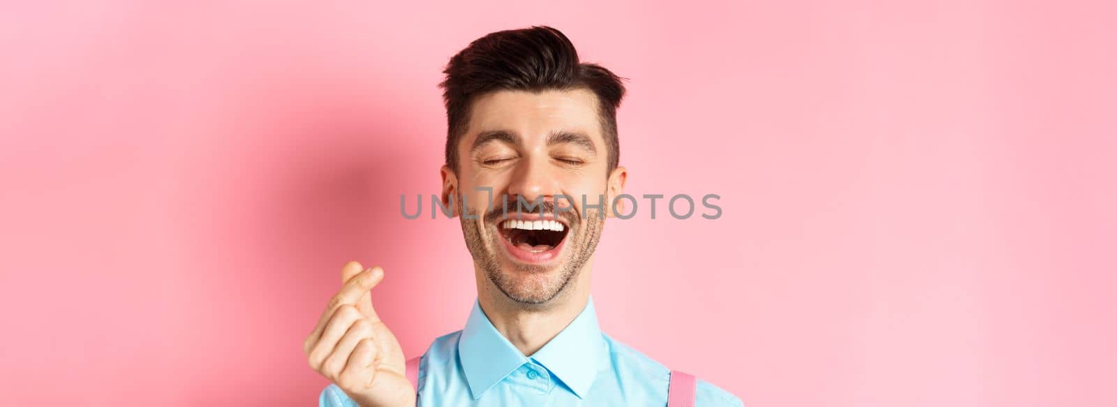 Valentines day concept. Happy man in love showing finger heart and laughing, standing on romantic pink background.