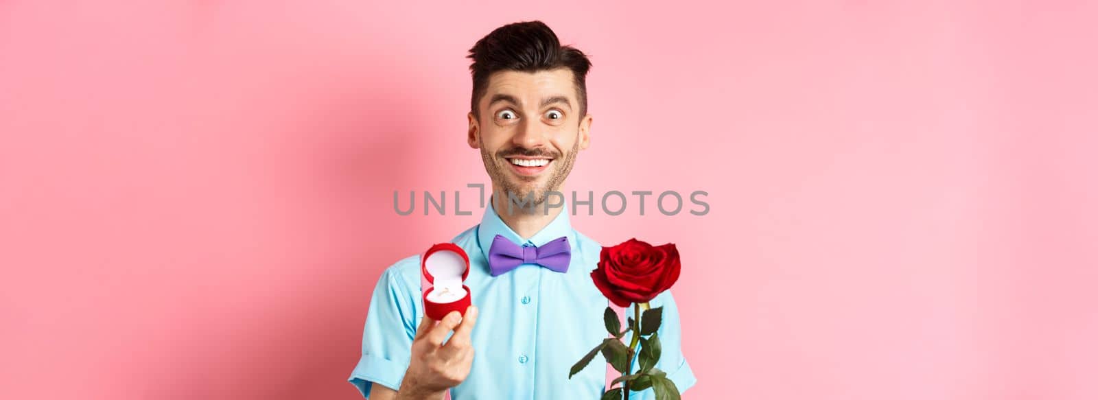 Valentines day. Smiling handsome man asking to marry him, showing engagement ring and red rose, standing romantic on pink background by Benzoix