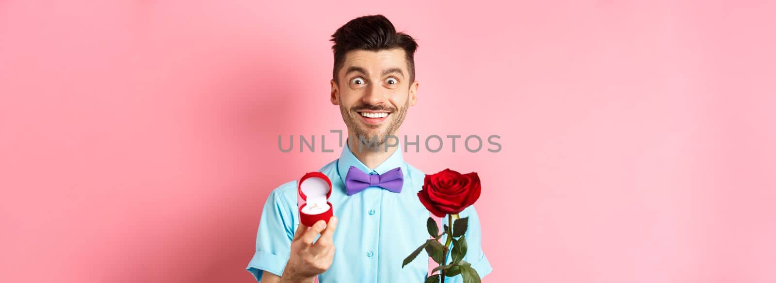 Valentines day. Smiling handsome man asking to marry him, showing engagement ring and red rose, standing romantic on pink background.