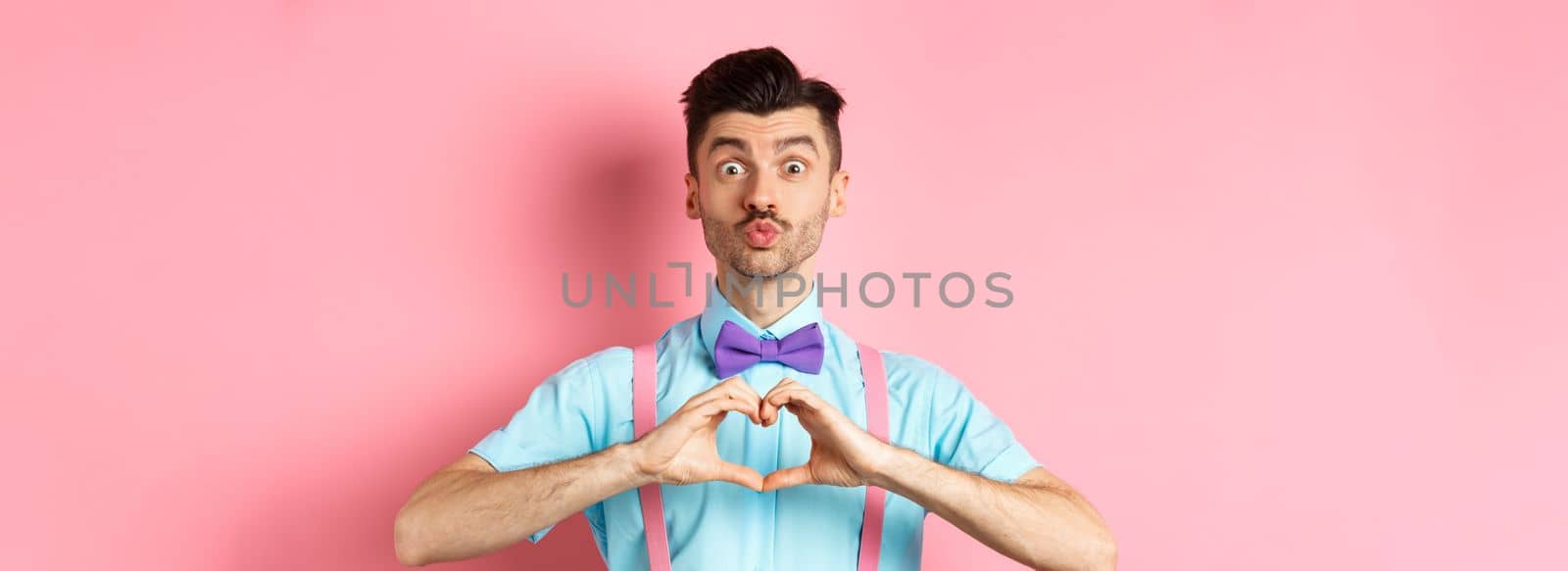 Happy Valentines day. Funny young man waiting for lovers kiss, pucker lips and show heart sign, I love you gesture, express feelings on date, pink background by Benzoix