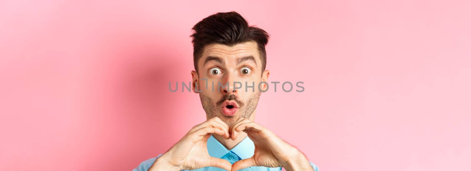 Valentines day concept. Close up of handsome caucasian man falling in love, looking amazed and showing heart gesture, pink background.