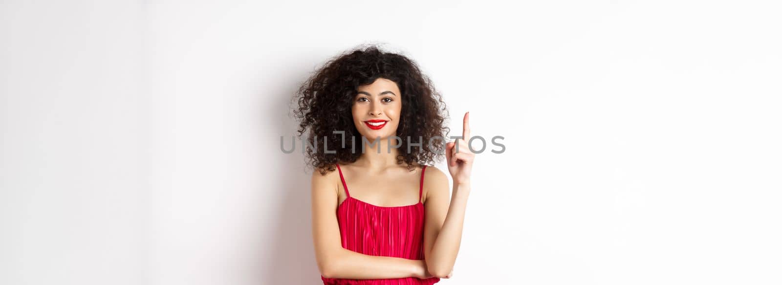 Beautiful smiling lady in red dress showing number one, raising finger and looking pleased, standing over white background.