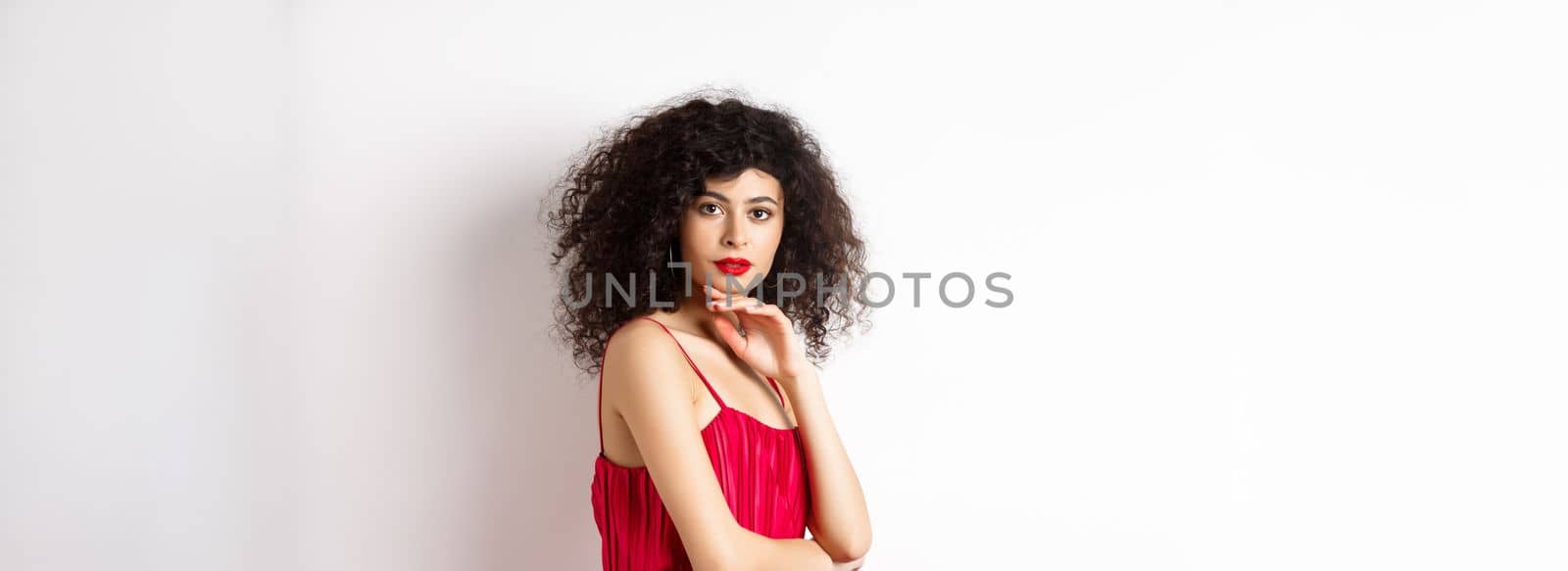 Beauty and fashion. Elegant lady with curly hair and red lips, fashionable makeup, wearing dress, gently touching chin and looking sensual at camera, white background.