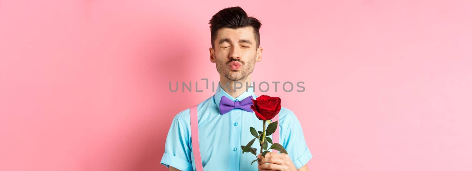 Cute and funny man waiting for kiss from lover on Valentines day, holding beautiful red rose for girlfriend, standing over pink background.