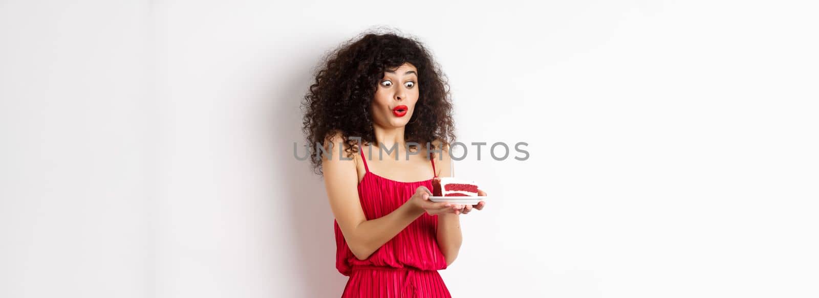 Excited birthday girl in red dress blowing candle on cake and making wish, standing on white background.
