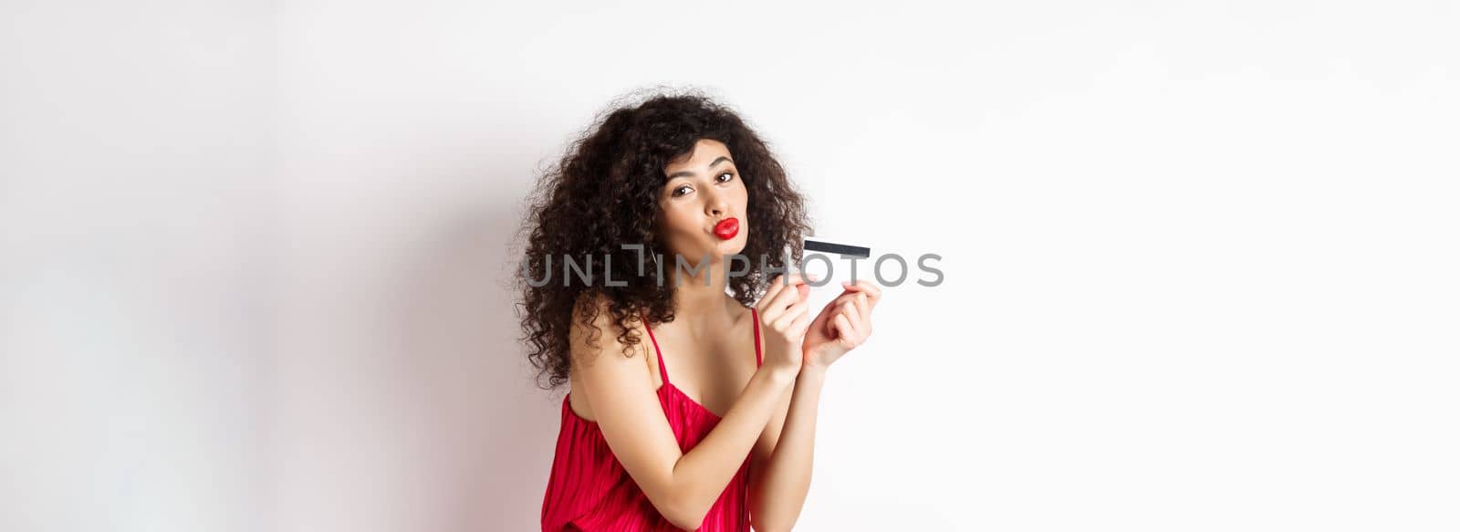 Shopping. Beautiful lady with curly hair, pucker lips, kissing plastic credit card, standing in red dress against white background.
