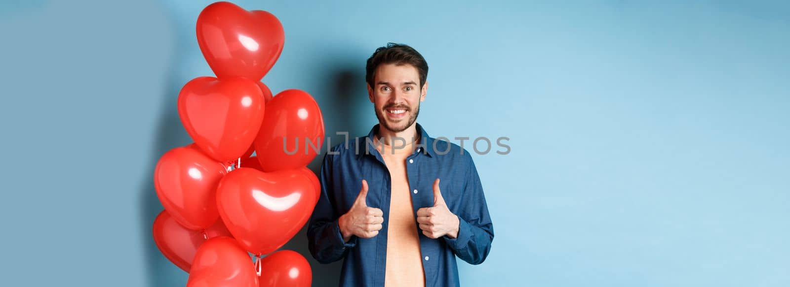 Happy valentines day. Cheerful boyfriend showing thumbs up in approval, standing with red hearts balloons for lover, blue background.