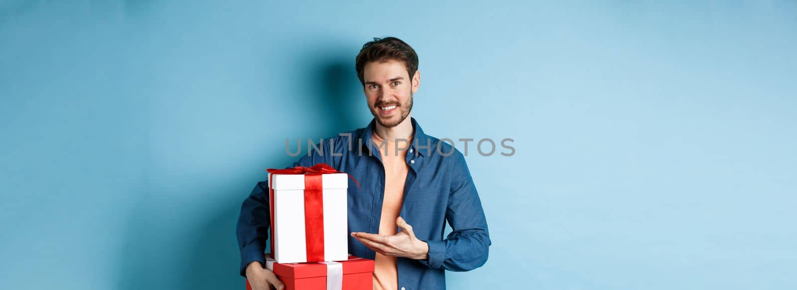Young man in casual clothes buying romantic gifts on Valentines day, pointing at presents boxes and smiling, standing over blue background by Benzoix