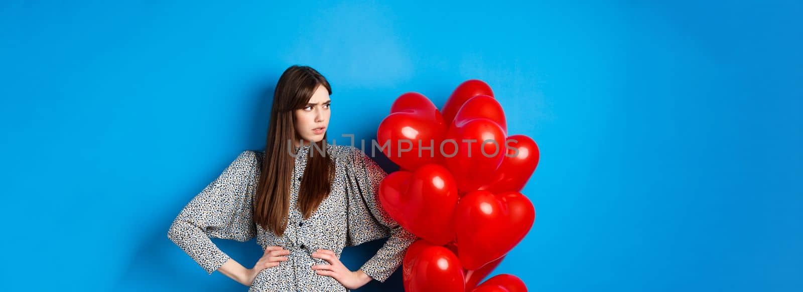 Valentines day. Annoyed and bothered girl complaining, looking left at empty space and frowning, standing near heart balloons, blue background.