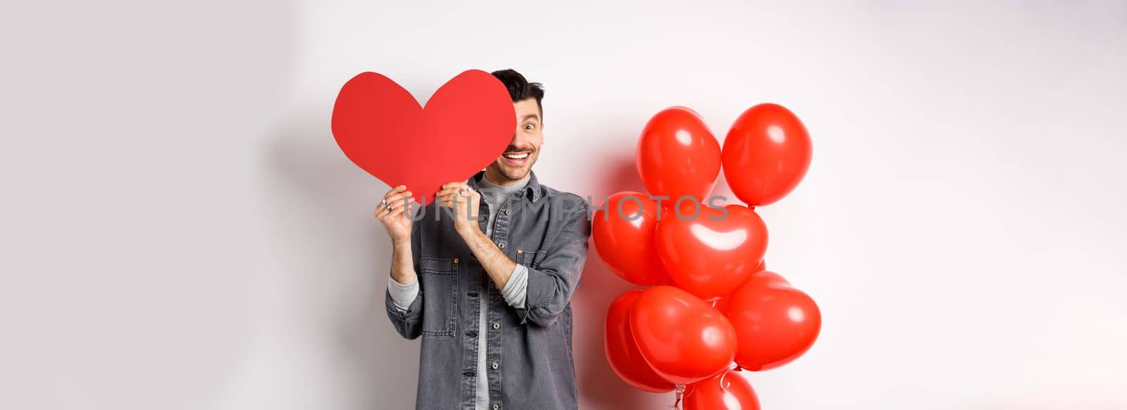 Romantic smiling man cover face with Valentine heart card and looking happy at camera, celebrating lovers day with partner, standing on white background.