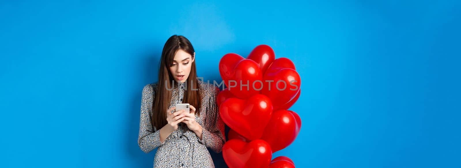 Valentines day. Portrait of young woman standing near red romantic balloons, looking surprised at smartphone screen, blue background.
