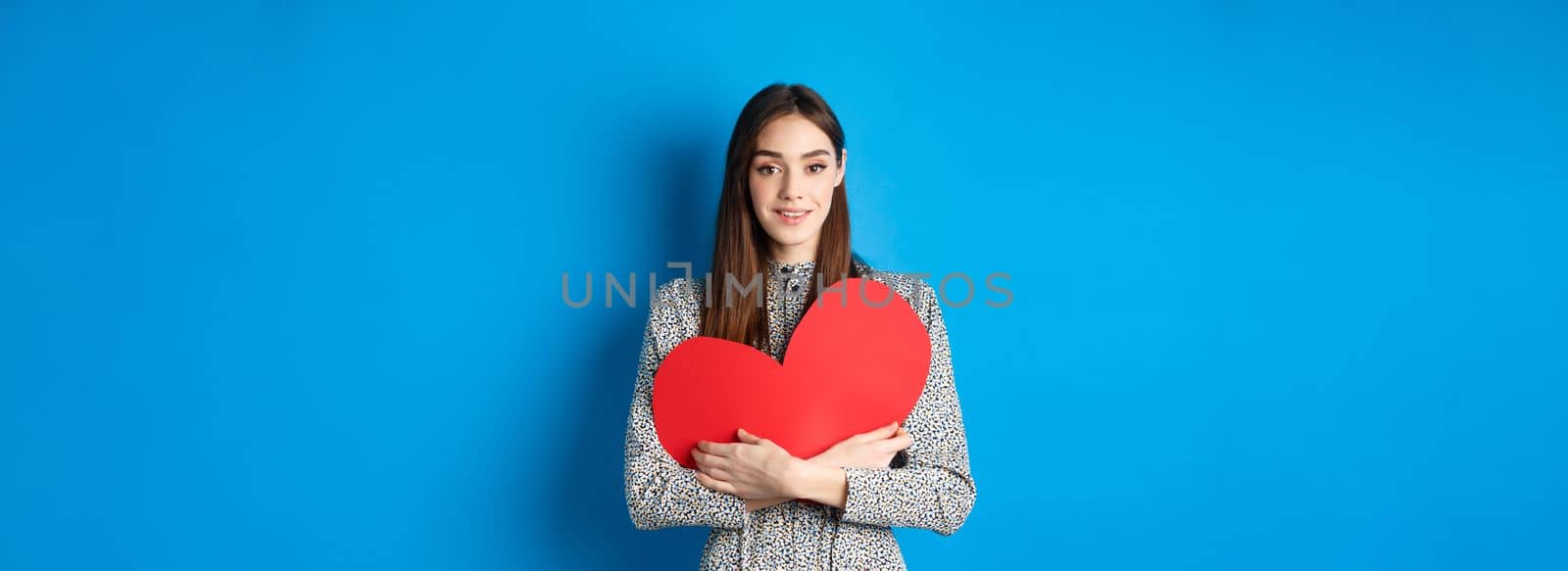 Valentines day. Attractive young woman searching for love, holding big red heart cutout and smiling at camera, standing in dress on blue background by Benzoix