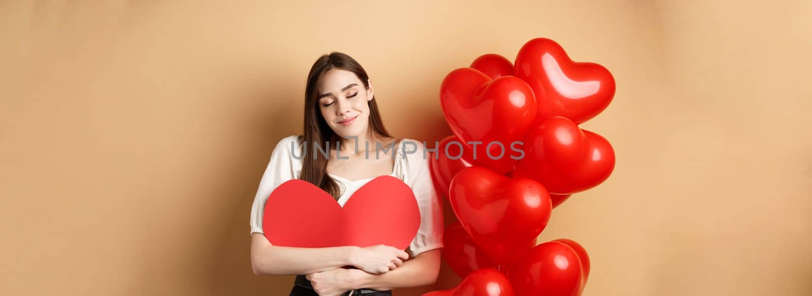 Romantic woman hugging big red heart and smiling dreamy, falling in love on Valentines day, dreaming about lover, standing on beige background near balloons.