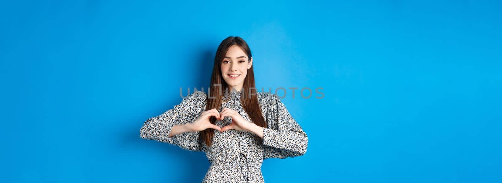 Pretty girl in romantic dress showing I love you heart gesture, smiling at camera, express sympathy and romance, standing on blue background by Benzoix