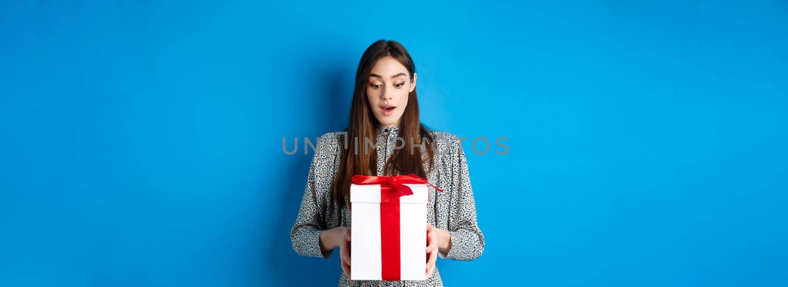 Holidays. Surprised young woman looking at gift with amazed face, receive Valentines day present, standing in dress on blue background.