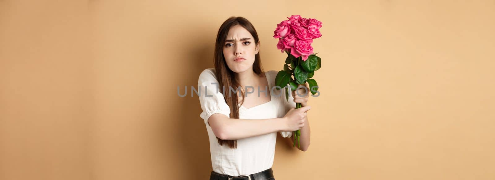 Happy valentines day. Angry girl hitting someone with flowers, swinging bouquet of roses at boyfriend, fighting with lover, standing on beige background by Benzoix